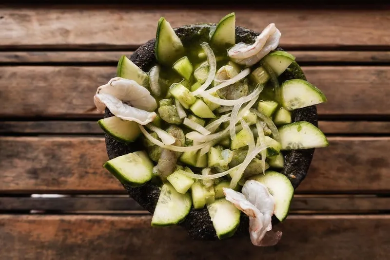 Aguachile negro — raw Sea of Cortez crystal shrimp cured in charred chile-lime sauce, served with sliced cucumber and red onion on a blue ceramic plate