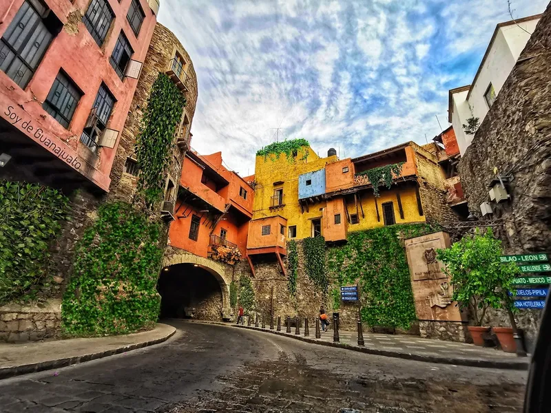 Narrow street beneath colorful hillside buildings, with stone walls, ivy, and dramatic clouds