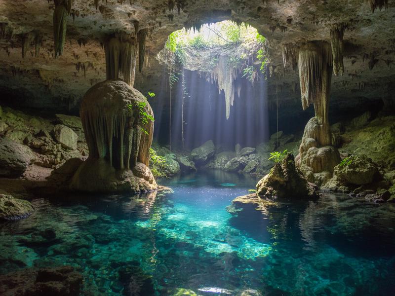 Underground cenote cave with dramatic stalactites hanging from the ceiling and light rays penetrating the clear blue water below